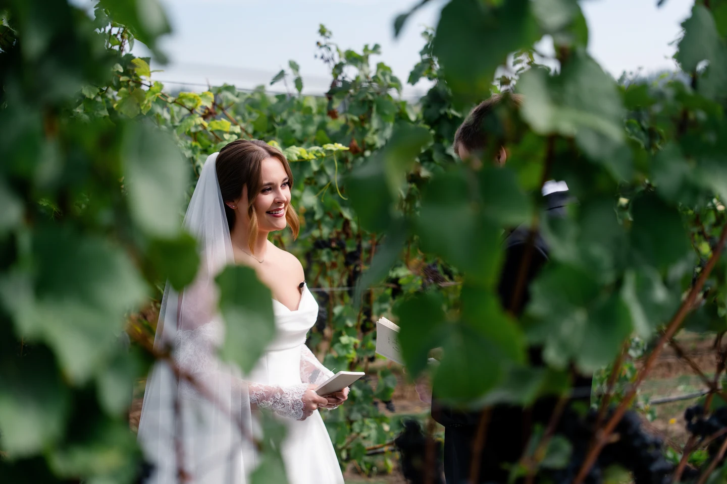 A bride smiles through vineyard leaves while holding her vows.