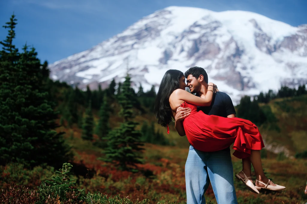 A couple embraces, with one partner lifted in a red dress, in front of a snow-covered mountain.