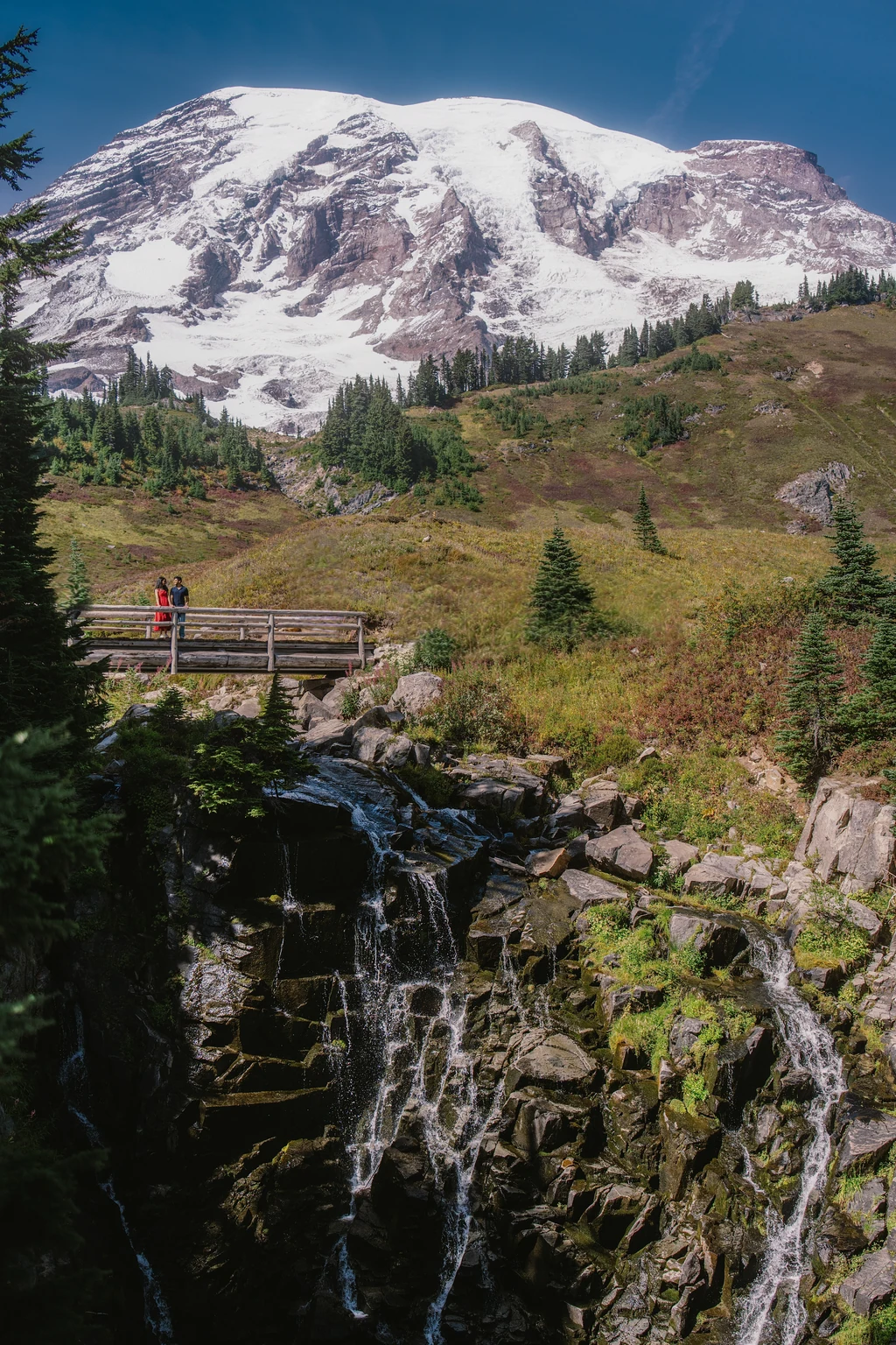 A couple stands on a wooden bridge above a waterfall, with a snow-covered mountain rising behind them.