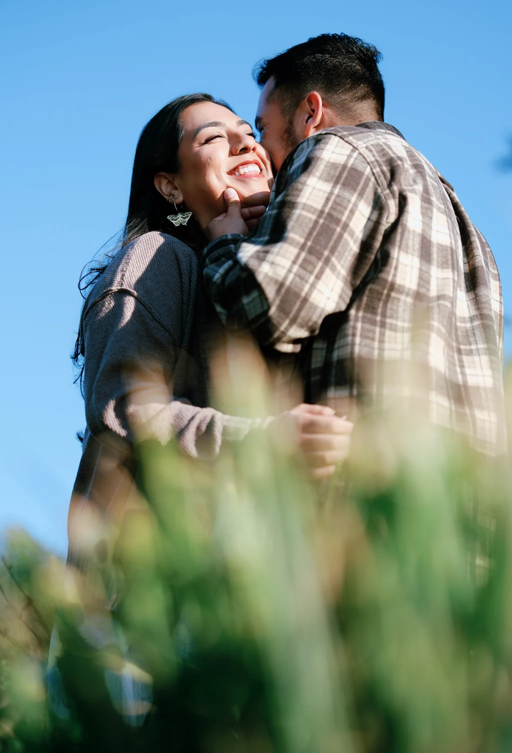 A smiling couple embraces against a clear blue sky, framed by softly blurred beach grass.