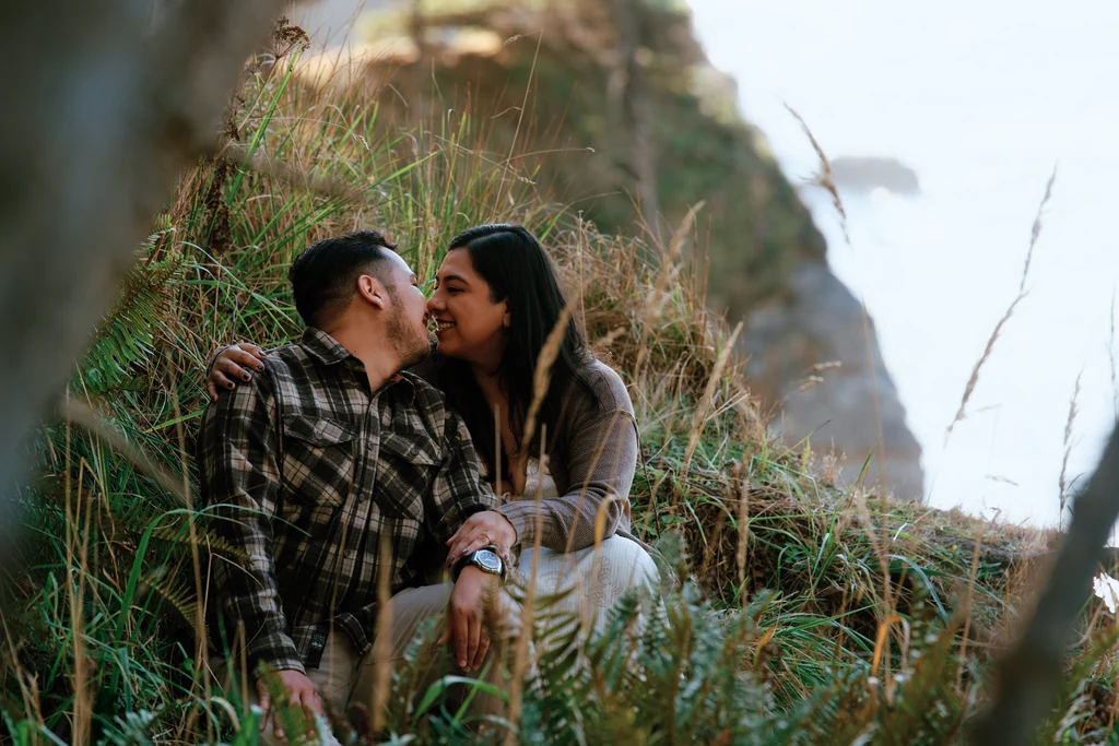 A couple shares a cheek kiss while sitting together in tall coastal grass beside a rocky cliff.