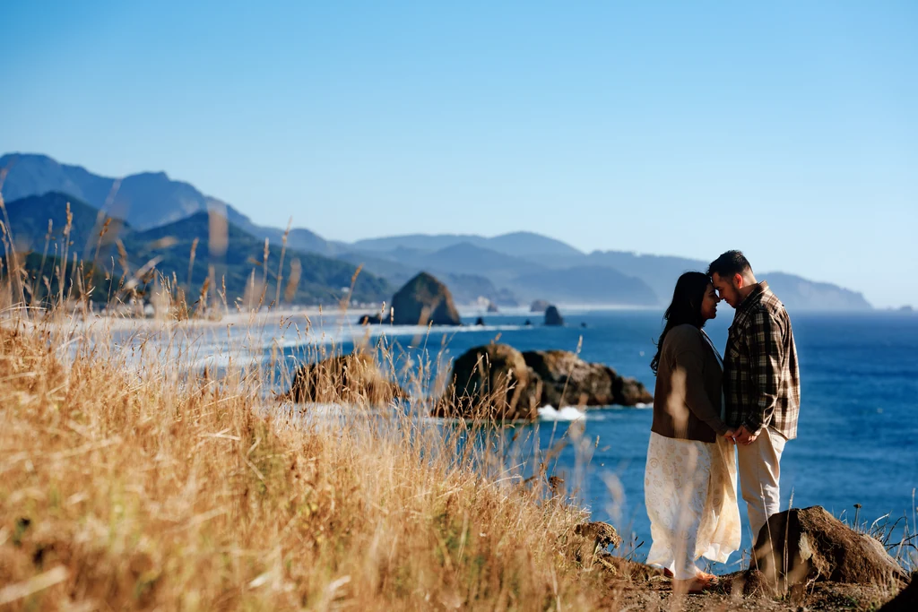 A couple shares a kiss on a grassy coastal bluff overlooking blue ocean water and offshore sea stacks.