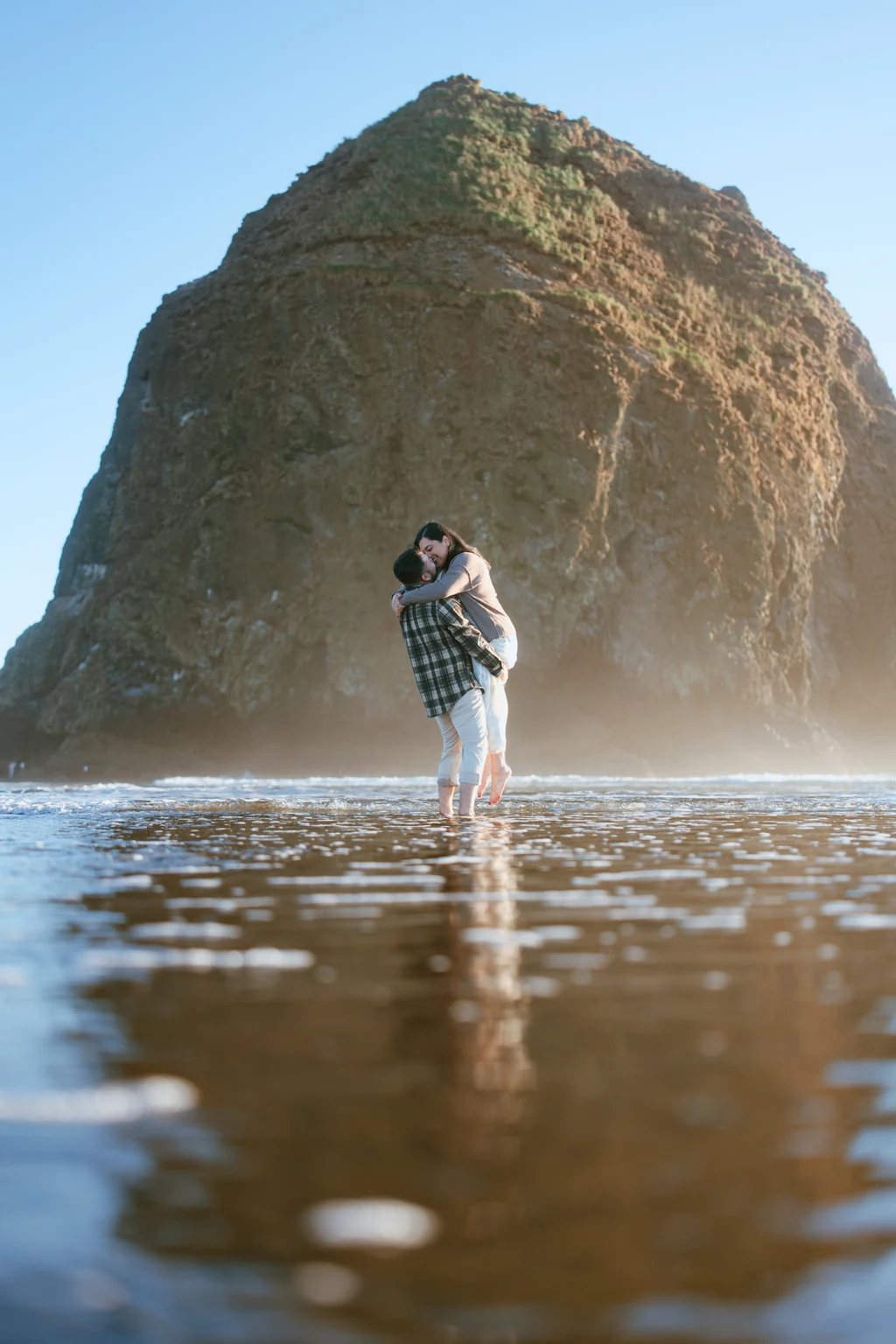 One partner lifts the other in shallow ocean water beneath a towering sea stack.