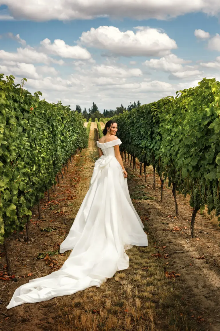 Wedding portrait of a person in an off-the-shoulder gown standing between vineyard rows beneath a cloud-filled sky.
