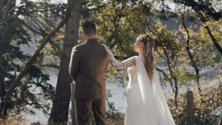 Newlyweds standing beside a wooded lake, viewed from behind.
