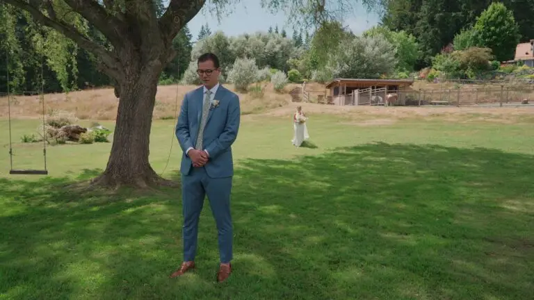 One newlywed waits beneath a tree as their partner approaches across a lawn.