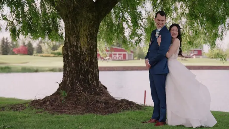 Newlyweds embracing beneath a tree beside a pond.